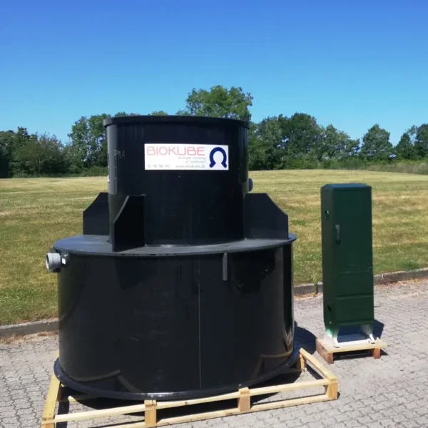 En stor svart cylindrisk minireningverk märkt "Biokube" är placerad på en träpall utomhus. Bredvid den finns en grön rektangulär struktur, en del av Biokube Venus Kombi-systemet. Bakgrunden presenterar ett gräsbevuxet fält och träd under en klarblå himmel.